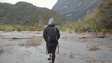 Girl-in-hiking-gear-walking-along-beach-surrounded-by-lush-green-forrest-and-mountains