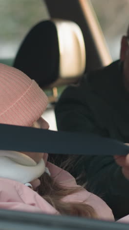 un hombre con una chaqueta verde está atento a atar el cinturón de seguridad de su hija pequeña, que lleva una chaqueta rosa y una gorra rosa, dentro de un coche. la escena captura un tierno momento de cuidado parental