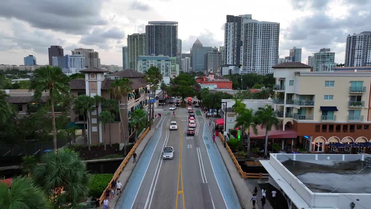 View of Las Olas Boulevard in downtown Fort Lauderdale, featuring high-rise buildings in the distance. Cars travel along the street, lined with palm trees and restaurants. Aerial.