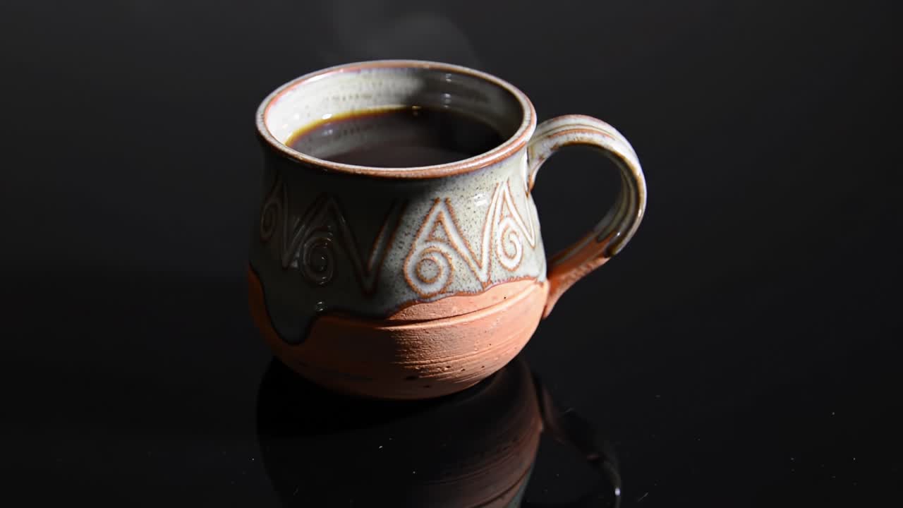 A beautifully crafted pottery mug filled with rich black coffee, emanating steam in a dark, reflective setting, showcasing the artistry of ceramic craftsmanship and beverage enjoyment