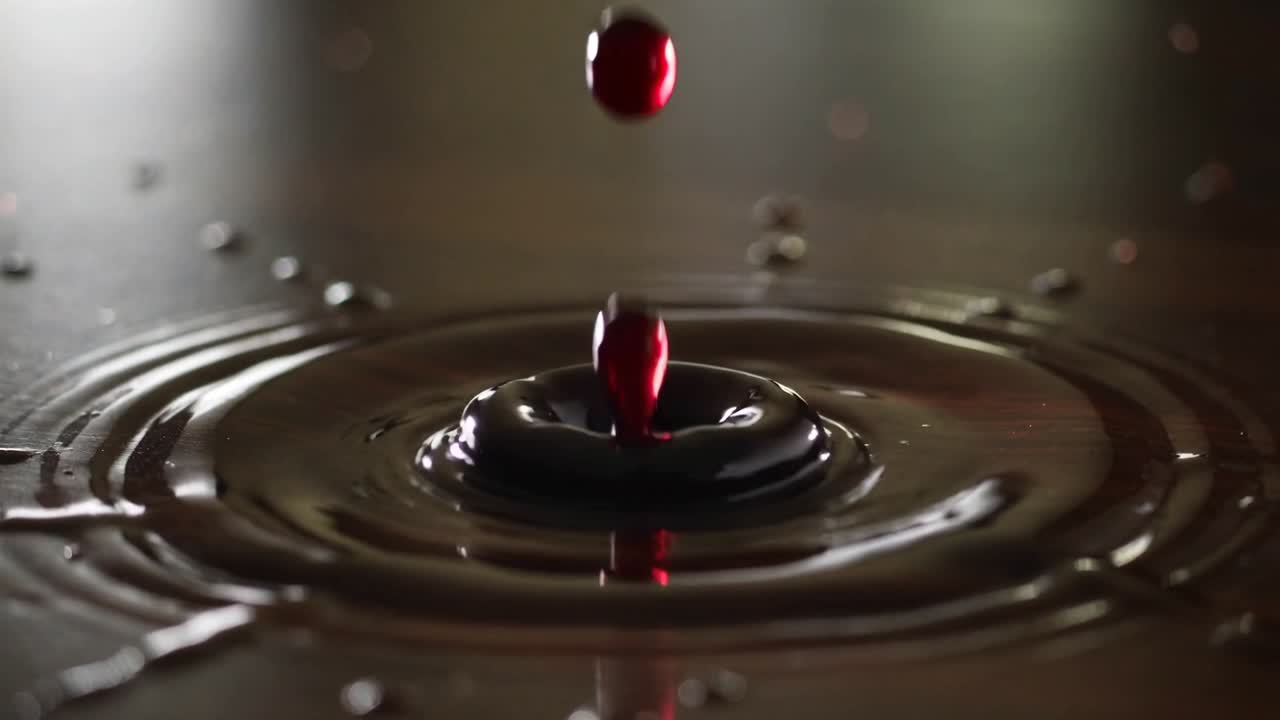 Goccia di sciroppo rosso che cade e colpisce un tavolo scuro e lucido, formando uno schizzo a corona, un getto di rimbalzo e satelliti.