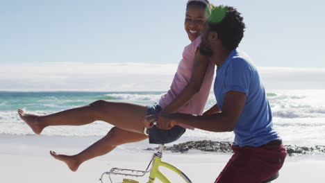african american couple riding on bike on the beach, woman sitting on handlebars