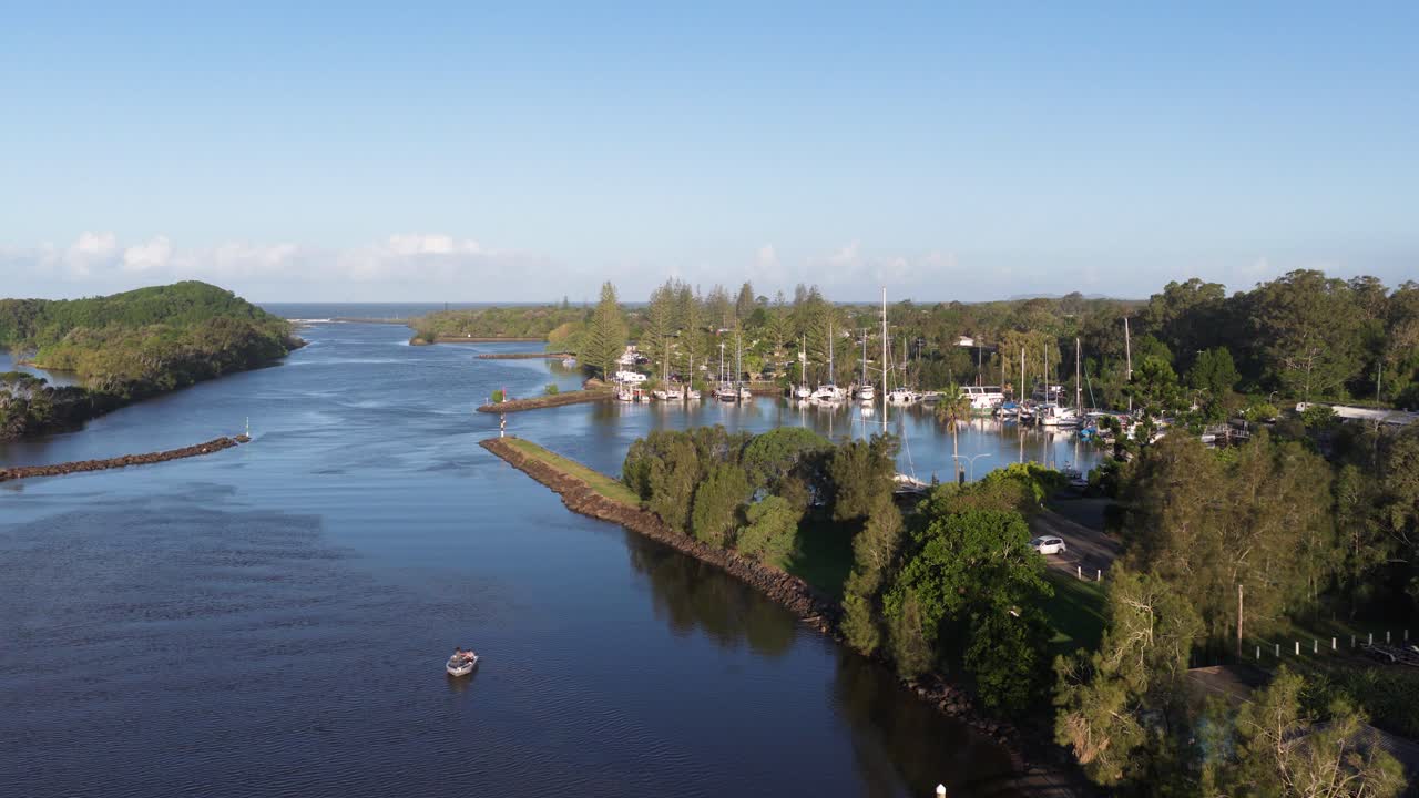 Drone footage captures serene river and marina at Brunswick Heads, NSW. Clear skies, lush greenery, and calm waters create a tranquil scene