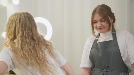 rear view of blond-haired customer sitting under ring light in beauty salon, gently swinging long hair backward as she settles in for hair or beauty session with soft lights illuminating background