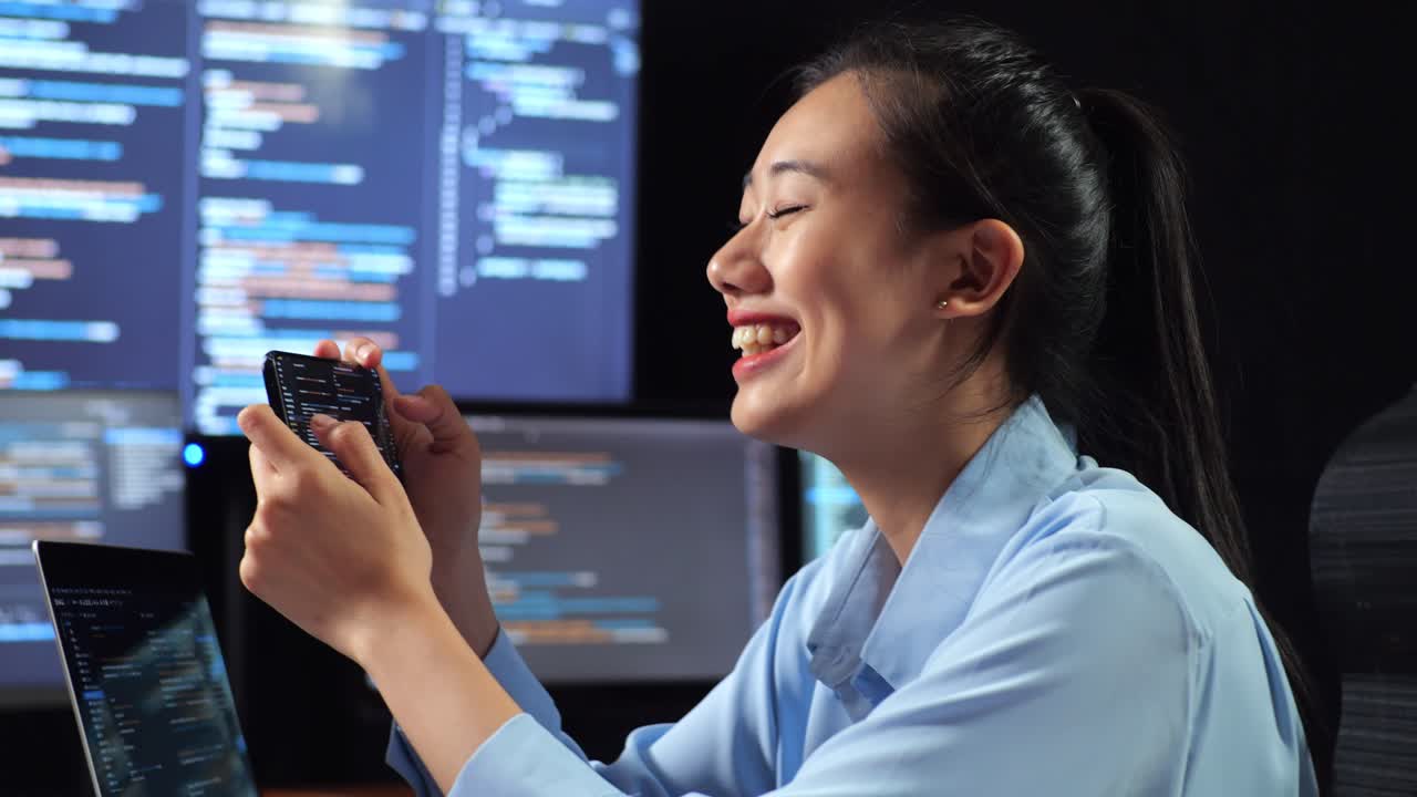 vista lateral de cerca de una programadora asiática usando un teléfono inteligente mientras escribe código en una computadora portátil usando múltiples monitores que muestran la base de datos en los escritorios de ventana de terminal en la oficina