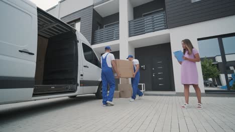 two young workers of removal company unload boxes and furniture from minibus into customer's home