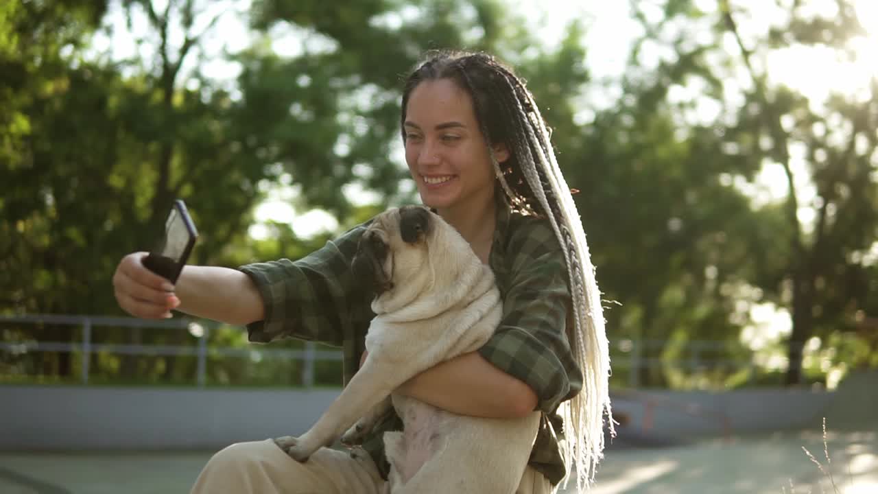 chica sonriente tomando una foto selfie con un lindo cachorro de pug en el parque de la ciudad verde sosteniendo un teléfono inteligente. bengalas de lente