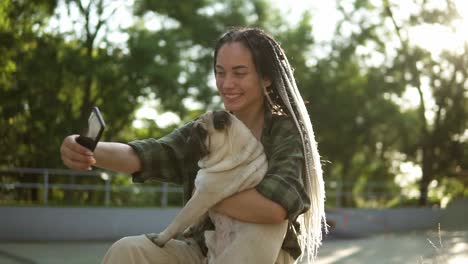 chica sonriente tomando una foto selfie con un lindo cachorro de pug en el parque de la ciudad verde sosteniendo un teléfono inteligente. bengalas de lente