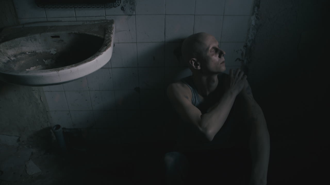 Pale bald skinny man sits next to the wall in the creepy old bathroom at night