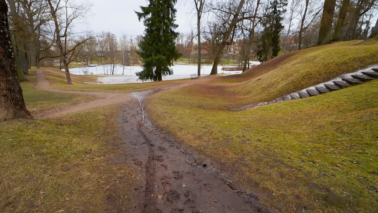 Mudret sti i en rolig park under forårets tøbrud, med smeltende sne, nøgne træer og en delvist frossen dam i baggrunden.