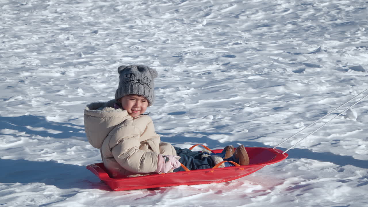 Glad smilende liten jente koser seg med å ake i snøen ved solnedgang, trukket av mamma - profilvisning, sporing