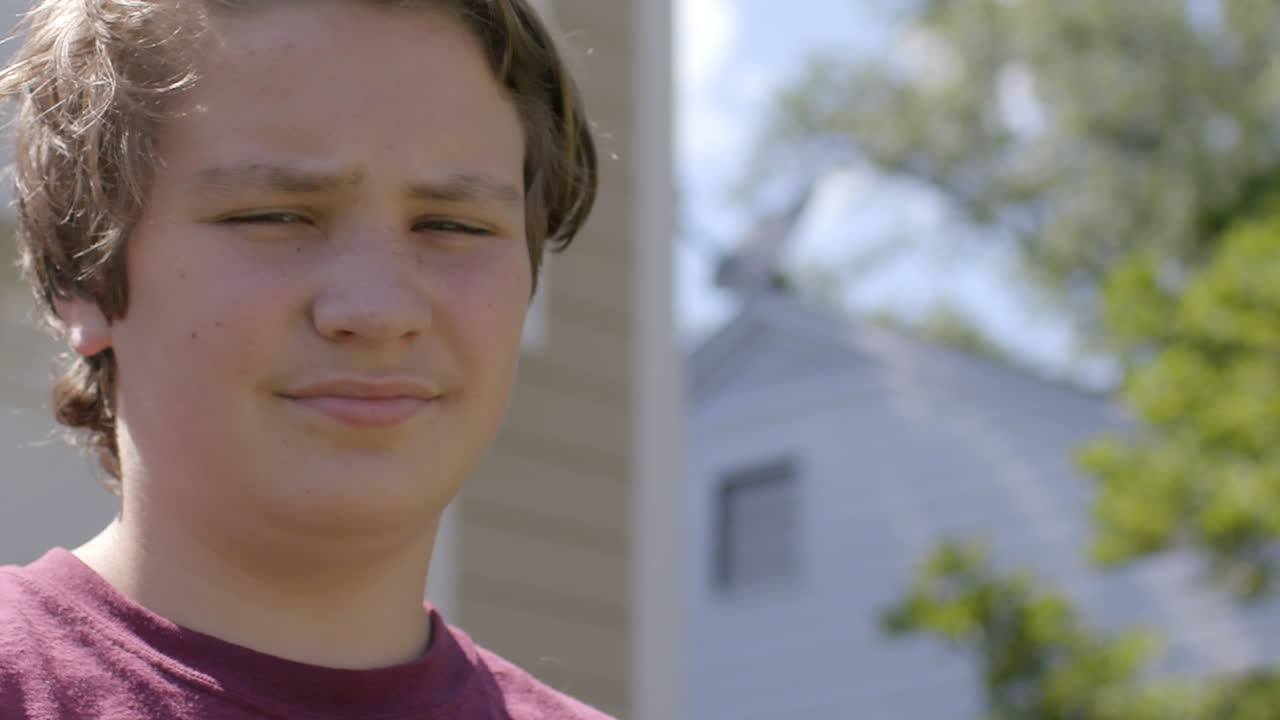 un adolescente frente a una casa suburbana en un día de verano mira la cámara