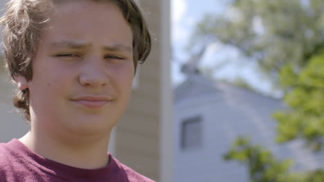 a teen boy in front of a suburban house on a summer day looks at camera