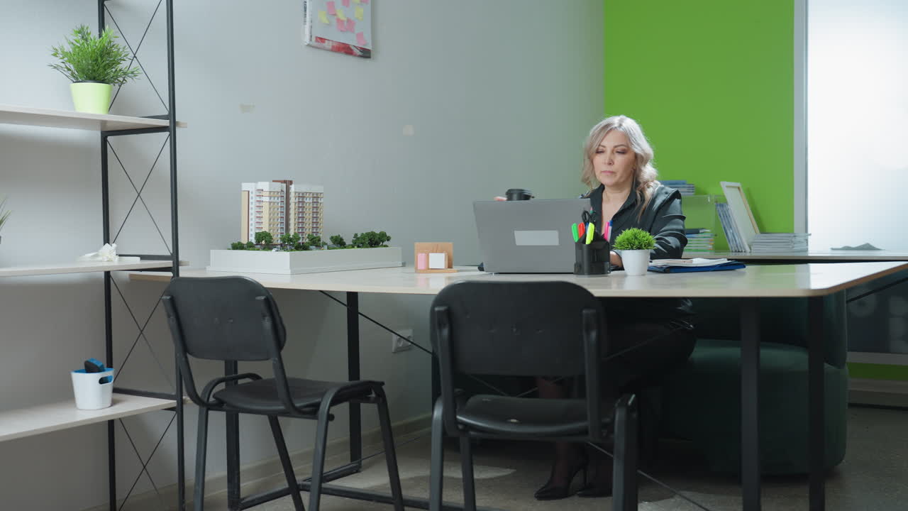 Real estate agent seated in modern office pauses work on laptop to take drink, sets cup aside, and resumes typing beside architectural model and organized desk items under natural light