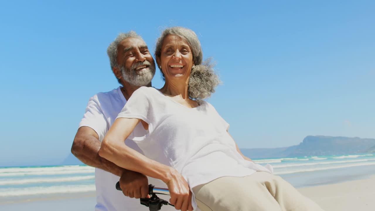 Front view of active senior African American woman sitting on man bicycle handlebars at beach 4k