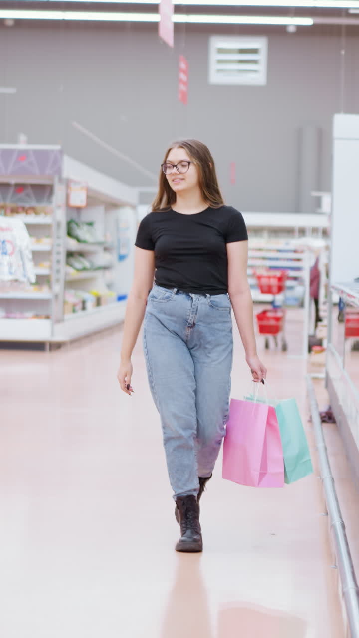 joven caminando por el centro comercial sosteniendo bolsas de compras, mirando la exhibición de vidrio con el reflejo visto en gabinetes elegantes, una señal colgada en el fondo y otra persona pasando en la lejanía