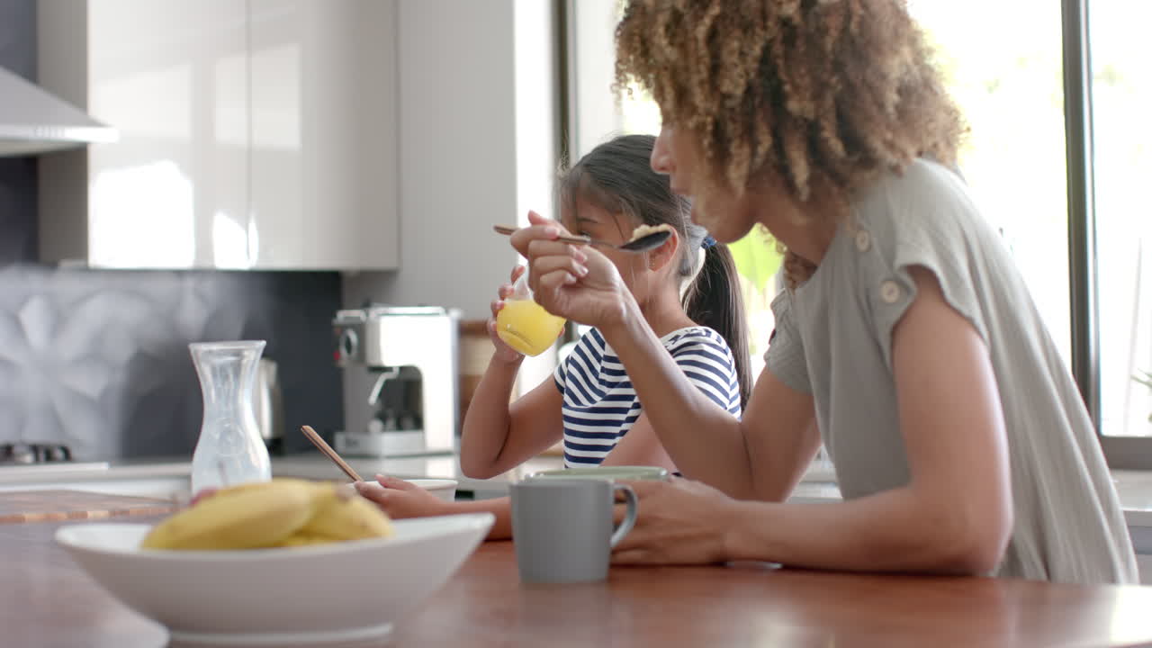 Madre e figlia multietniche che mangiano cereali a colazione in una cucina soleggiata, spazio per il testo, al rallentatore