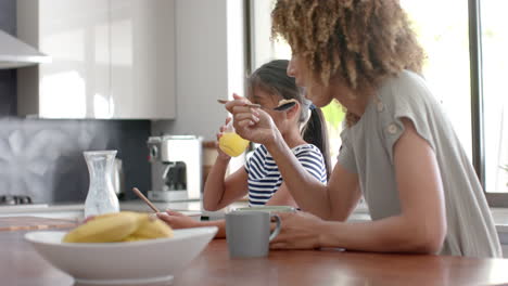 biracial mother and daughter eating breakfast cereal in sunny kitchen, copy space, slow motion