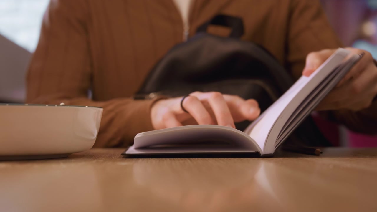 Primo piano di una persona con abiti marroni che apre e sfoglia le pagine di un quaderno su un tavolo di legno con illuminazione soffusa. Uno zaino nero e un portapenne sono visibili mentre il soggetto interagisce