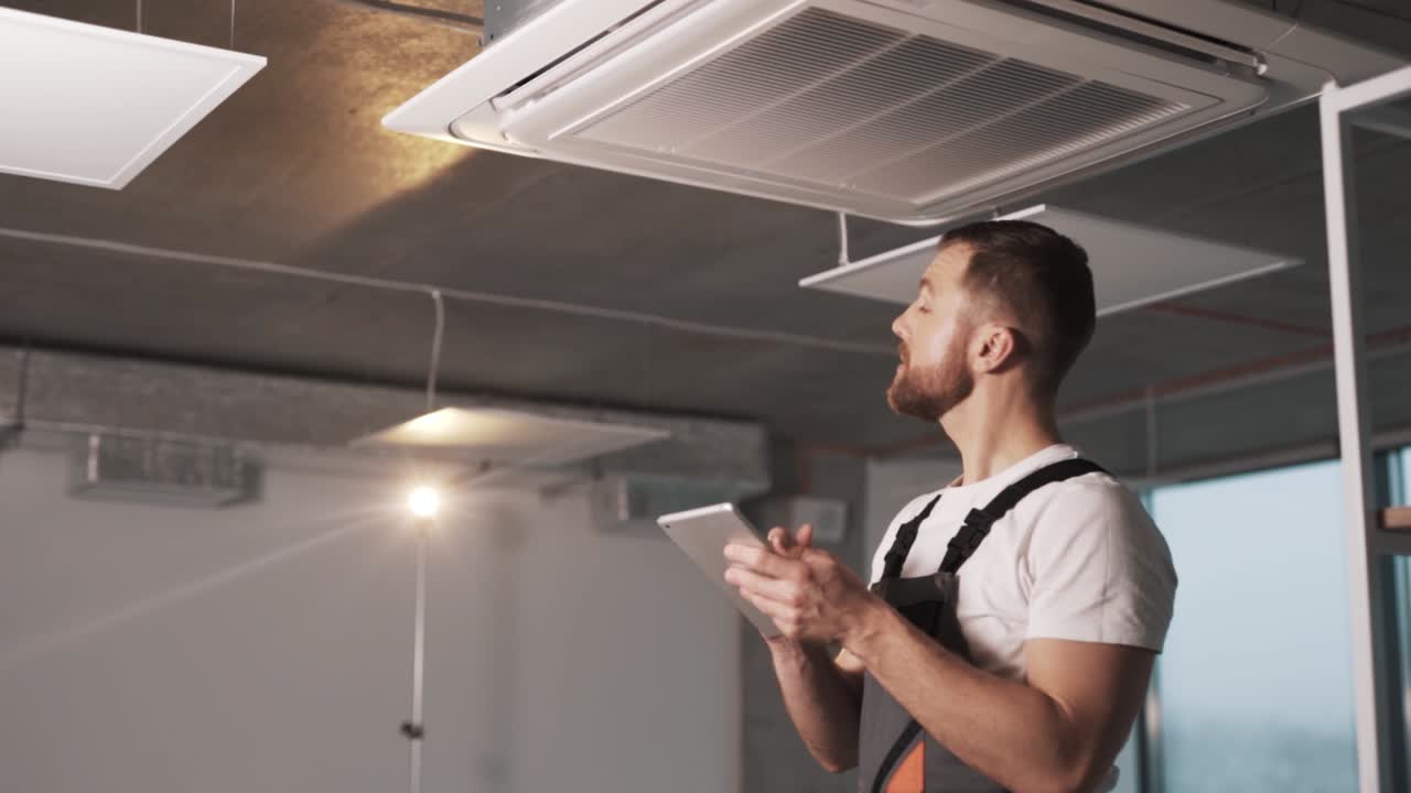 portrait of an adult man in a work uniform installing an industrial air conditioner and configuring software in a modern office