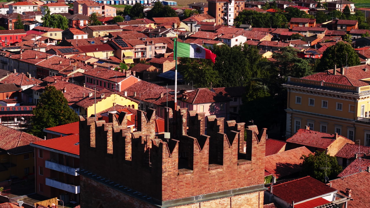 Drone shot tilting down while focused on the Italian flag flying above a medieval stone tower in a Piedmont town.