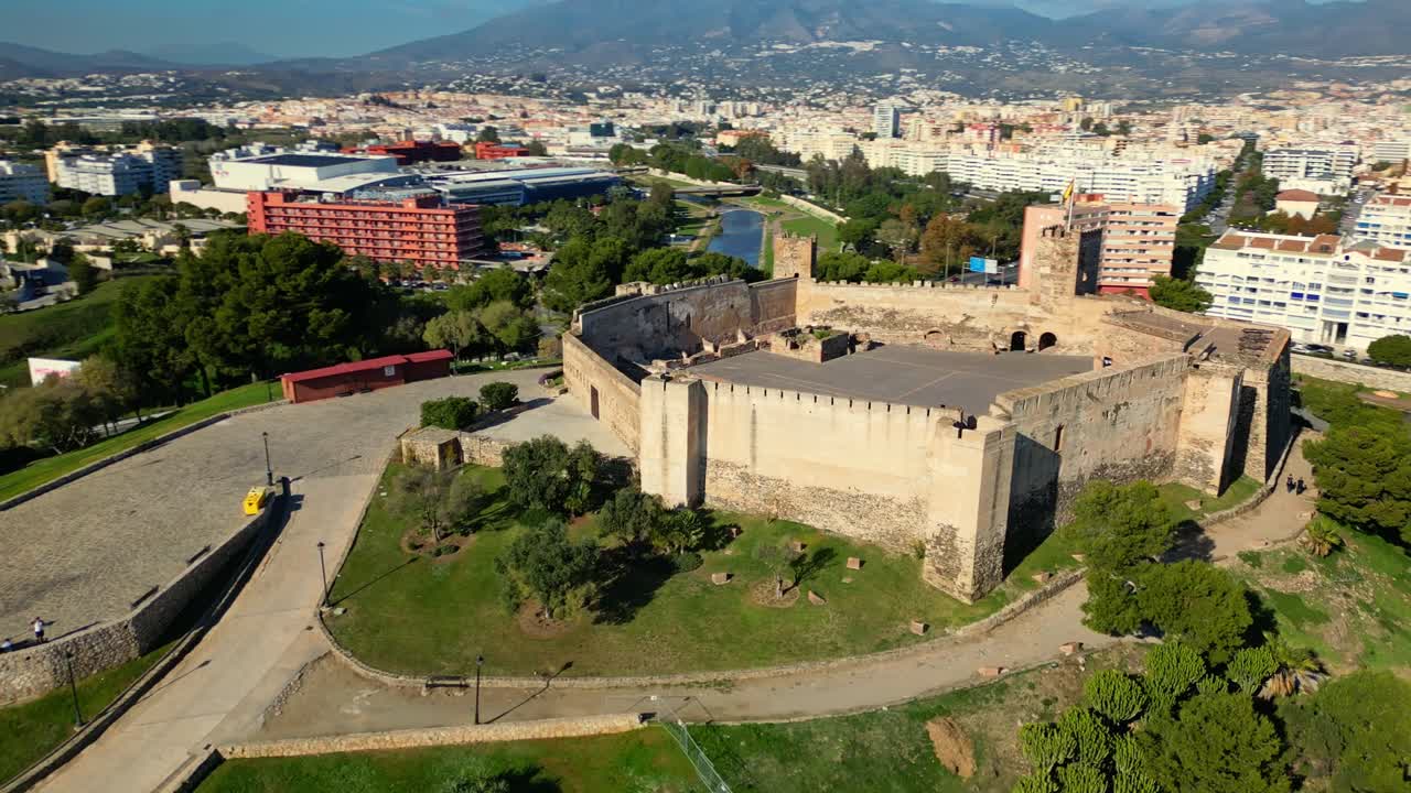 Orbita aerea del Castillo Sohail a Fuengirola, Costa del Sol, Spagna, con la bandiera spagnola che sventola sopra la storica fortezza mentre la vista si estende dalla costa mediterranea alla graziosa città costiera.