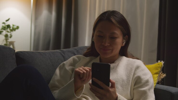 Woman Spending Evening At Home On Sofa Wearing Wireless Earbuds And Streaming Music Or Podcast From Mobile Phone 4