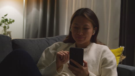 woman spending evening at home on sofa wearing wireless earbuds and streaming music or podcast from mobile phone 4