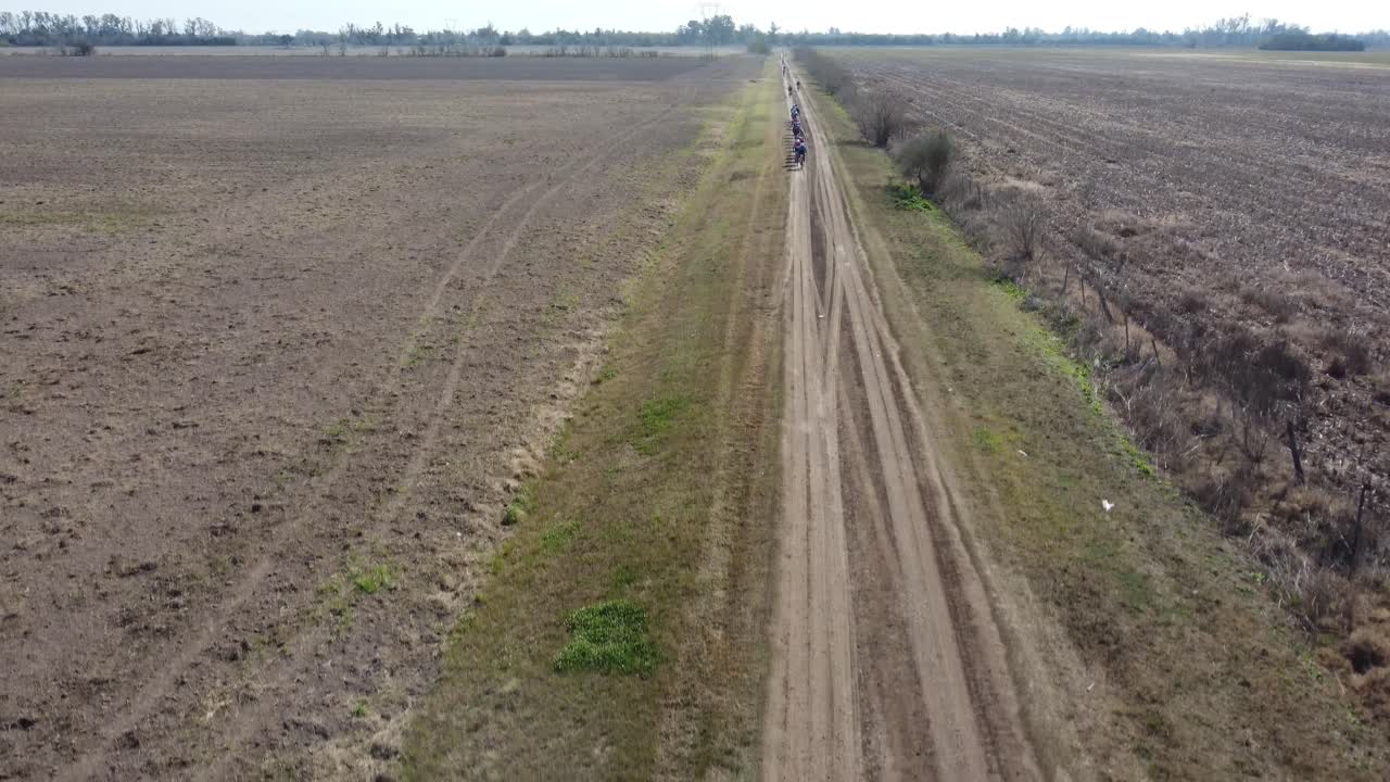 Grupp cyklister cyklar längs en landsväg förbi ett stort fält under molnig himmel