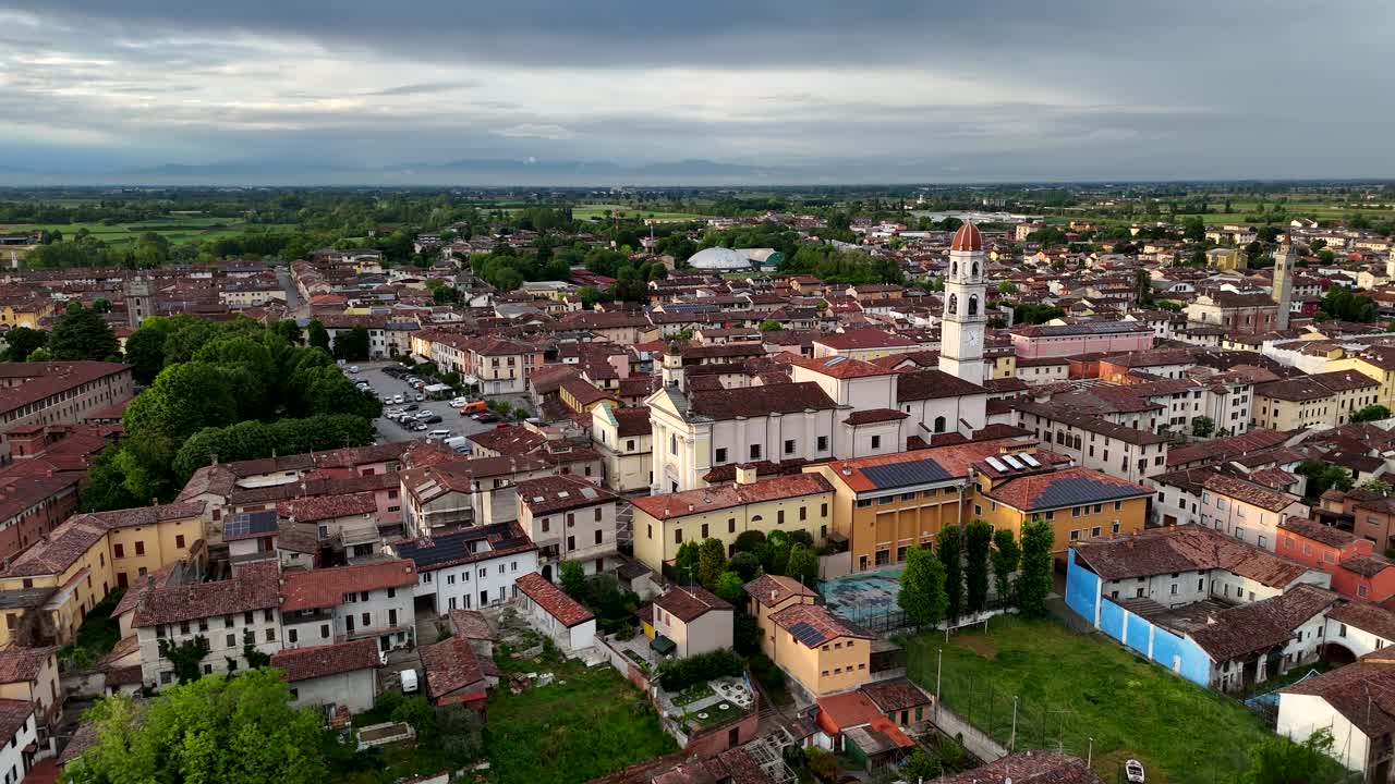 Drone orbit captures the historic Church of Santi Tommaso e Andrea Apostoli standing prominently in Pontevico, Brescia in Lombardy, near red-roofed houses and the flat northern Italian plain at dusk