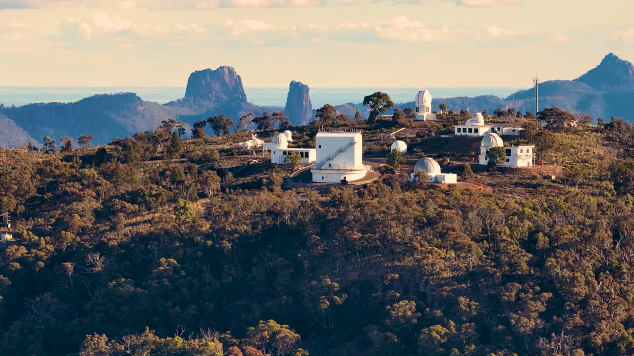 Aerial footage circles a mountaintop observatory complex with multiple telescopes, warm sunset lighting, and distant mountain ranges in Coonabarabran, Australia