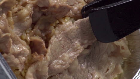 vista superior de rebanadas de carne cocidas añadidas y superpuestas sobre la superficie de arroz amarillo con pinzas de plástico negras en una bandeja de extracción de lámina plateada, directamente encima, detalle estático de primer plano extremo