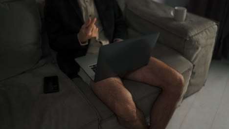 close-up a brunette man with stubble in a black jacket and beige shorts enters a video conference using a gray laptop. sitting on a sofa in a modern apartment working remotely in the evening