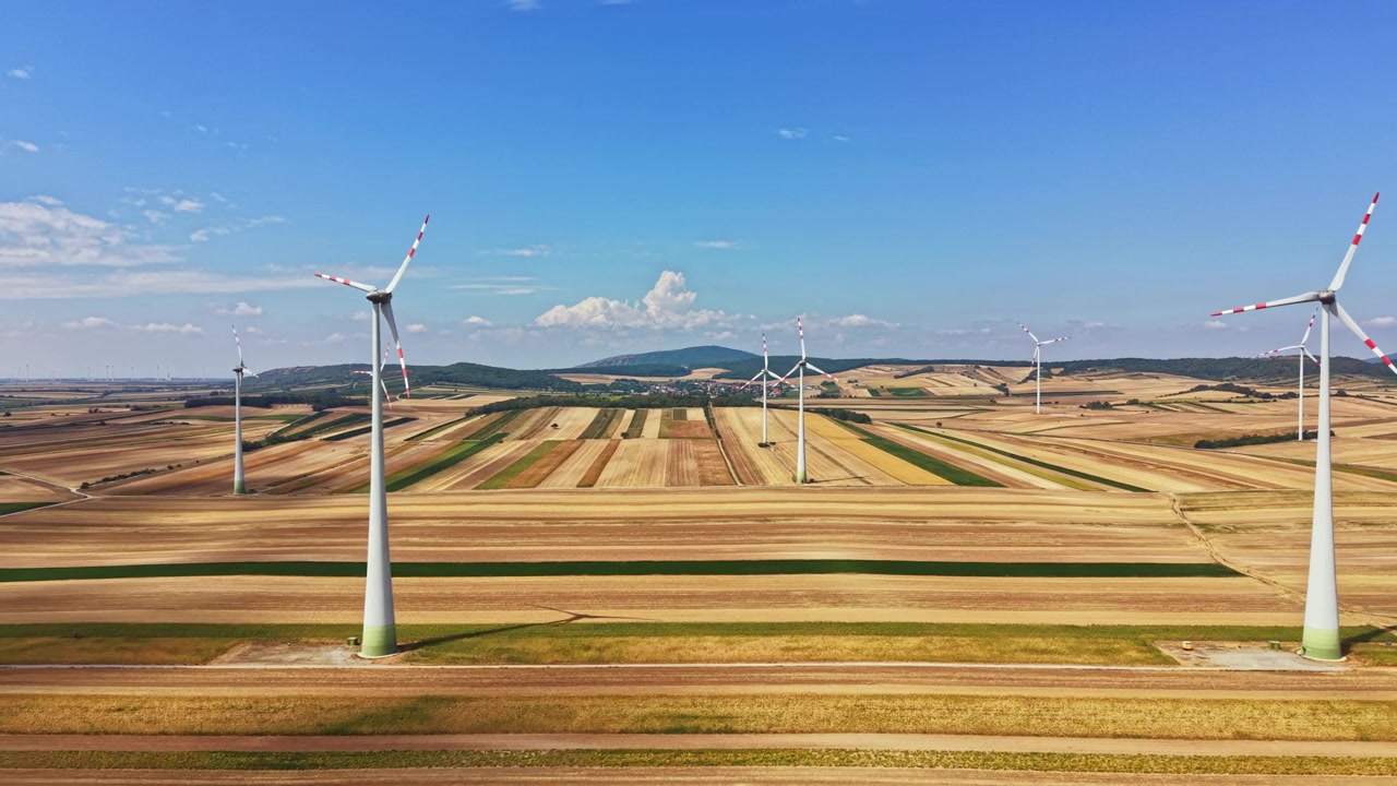 Wind turbines stand in a wide field during the day with clear skies and rolling hills in the background