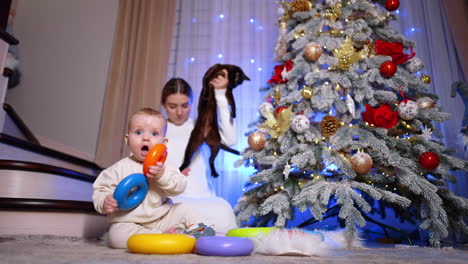 adorable baby sits at christmas tree playing with toys. woman sitting behind takes a black cat but the pet walks away. low angle view.