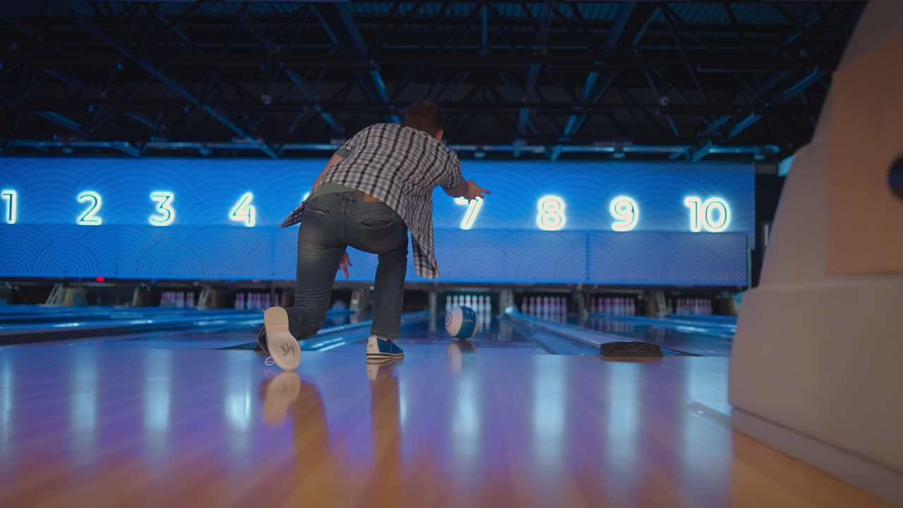 View from the back a man throws a ball and the camera follows him in a bowling club