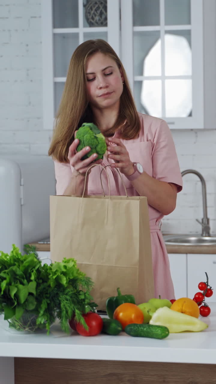 Young woman unpacking shopping bag, taking out green broccoli. Kitchen background and pretty girl putting fresh vegetables on the table. Healthy food. Vertical video