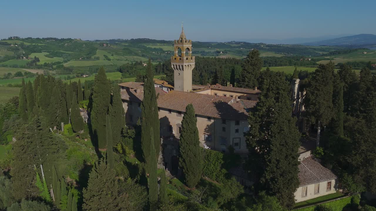 Drone shot approaching Castello di Montegufoni in Tuscany. Surrounded by cypress trees and vineyards, the castle emerges beautifully in the lush green Tuscan landscape under clear morning skies.