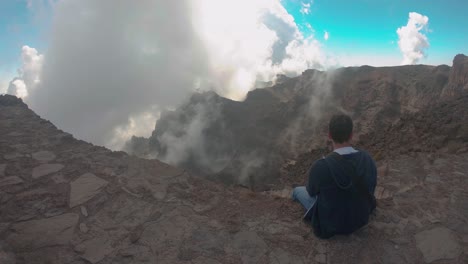 slow motion shot of a man sitting on a mountain and fog or mist rises slowly, between mountains, on a sunny day, in montenegro