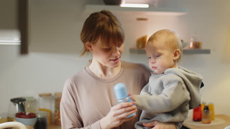 Mom Holding Baby Playing with Water Bottle in Kitchen