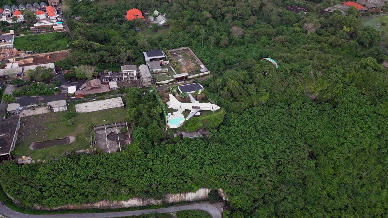 Aerial capture shows an airplane villa on a Bali green hill with an infinity pool facing the ocean and paragliding along the coast giving a view of this luxury stays above tropical landscape