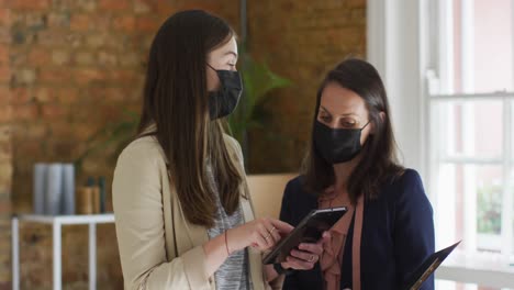 Caucasian-female-business-owner-and-her-coworker-wearing-face-masks,-using-tablet-and-talking