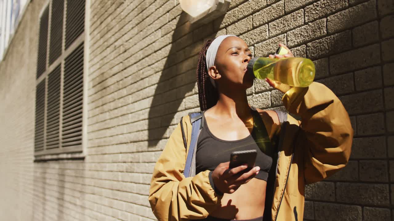 African american woman exercising outdoors drinking water and using smartphone in the city