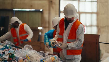 Un-Hombre-Moreno-Con-Barba,-Con-Uniforme-De-Protección-Blanco-Y-Chaleco-Naranja,-Junto-Con-Sus-Compañeros-Clasifica-Las-Botellas-De-Plástico-Por-Color-En-Una-Planta-De-Reciclaje-De-Residuos.