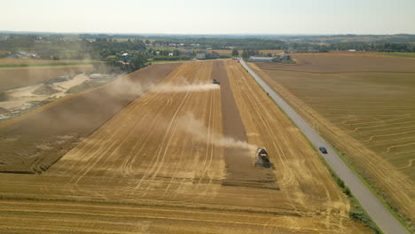 aerial backward flight showing harvesting machines on wheat field in middle of dust beside road with driving car