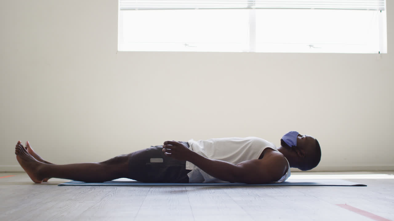 hombre afroamericano en forma que usa una máscara practicando yoga mientras está acostado en un estudio de yoga