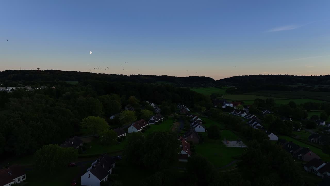 해가 지는 빛으로 조명된 새 무리가 아래의 정착촌 위를 날아다닙니다.