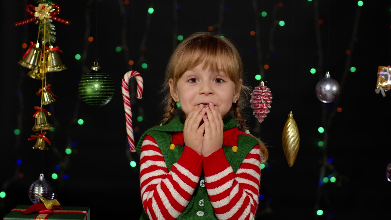 Holčička v kostýmu vánočního skřítka pomocníka Santa Clause se dívá do kamery, dělá velké oči a zakrývá si ústa.