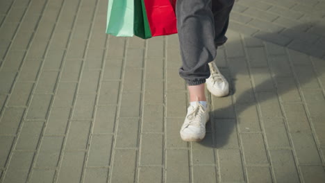 leg view of individual in grey joggers and white canvas shoes, holding green, mint, and red shopping bags while walking casually on an interlocked path walk pass a small piece of paper on the floor