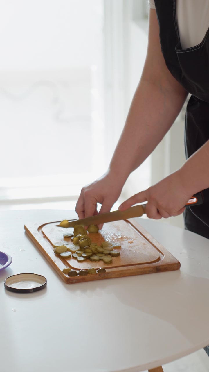 Primo piano di mani che affettano cetriolini a rondelle sottili su un tagliere di legno su un bancone luminoso della cucina, mostrando la preparazione di cibo fresco, un movimento attento del coltello, una superficie pulita e un dettaglio della cucina casalinga quotidiana.